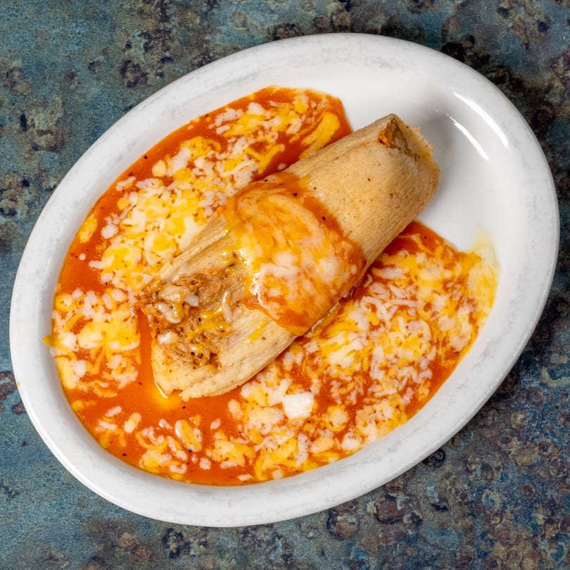 A single tamale covered in red sauce and melted cheese on a white oval plate.