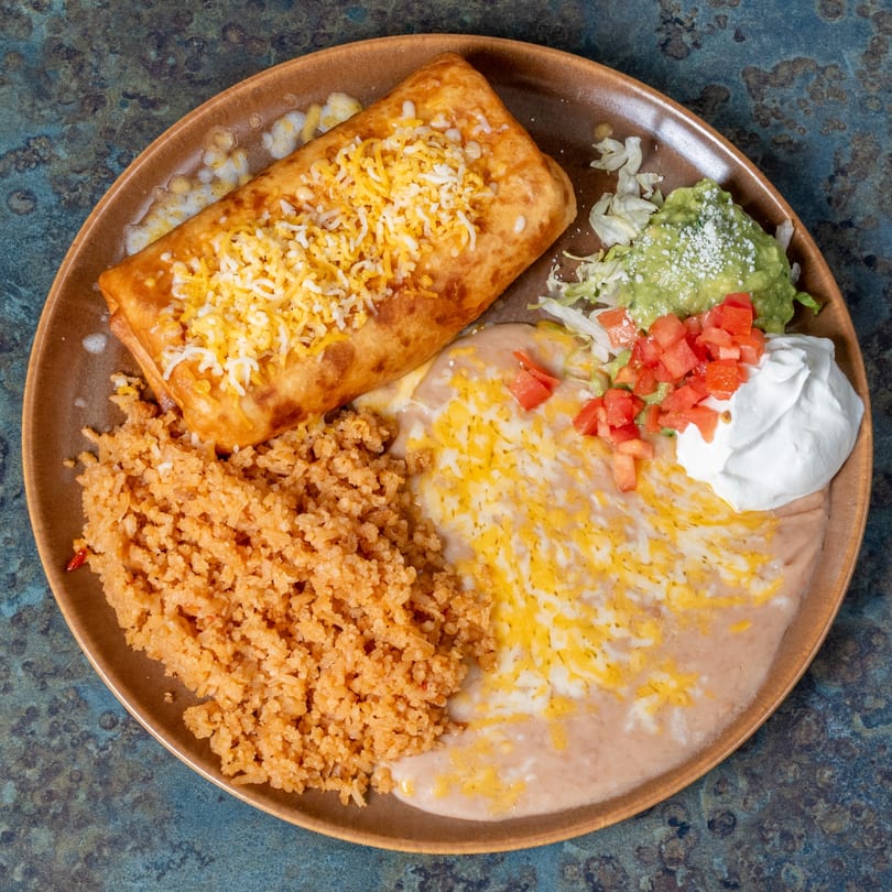A golden-brown chimichanga topped with cheese, served with rice, refried beans, guacamole, and sour cream.