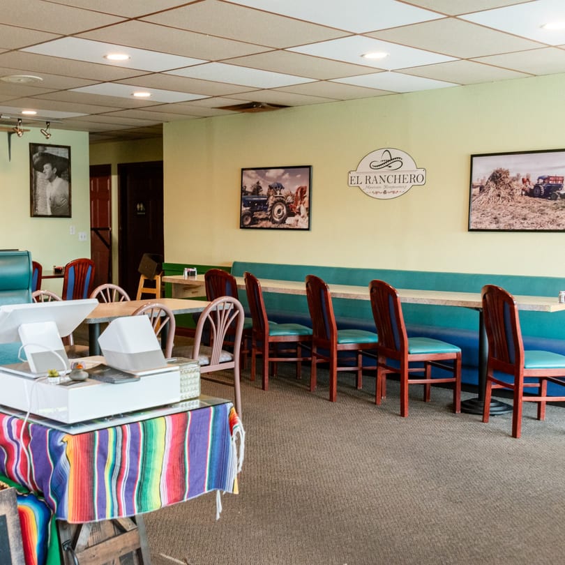 Interior of El Ranchero Mexican restaurant with colorful chairs, booths, and framed artwork on light green walls.