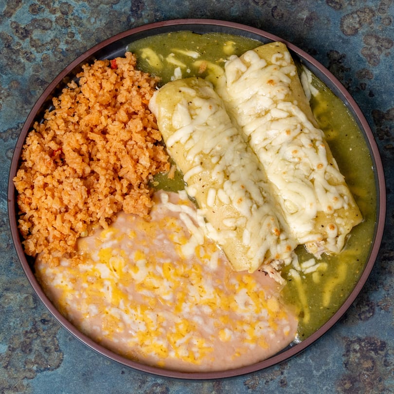 Two green enchiladas with melted cheese, served with Mexican rice and refried beans on a dark plate.
