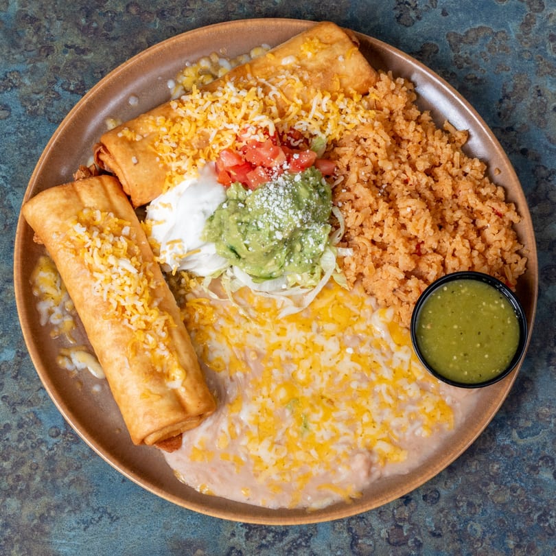 Two golden-brown flautas topped with cheese, served with rice, refried beans, guacamole, sour cream, and salsa.