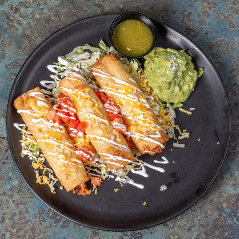 Three golden-brown El Ranchero taquitos topped with cheese, tomatoes, and sour cream, served with guacamole and salsa.