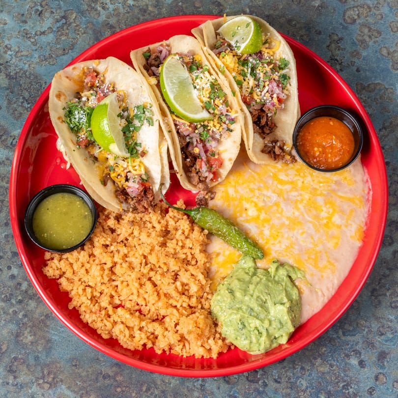 Three El Ranchero Mexican tacos with rice, beans, guacamole, and salsas on a red plate.